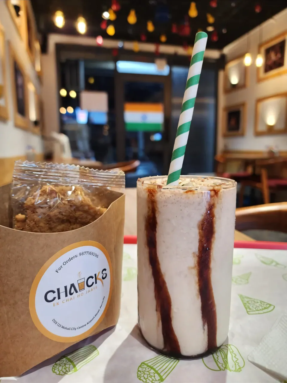 A chocolate-streaked shake with a green striped straw next to a CHAICKS kraft snack pack, inside the café